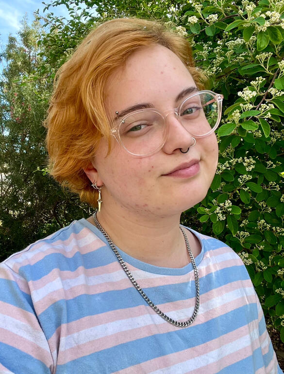 An old image of Riley, with his hair still grown out a few inches. His glasses are tilted downwards, looking at something. We see him through the gap between two bookshelves, with a dozen books in a row sat in the foreground of the image.