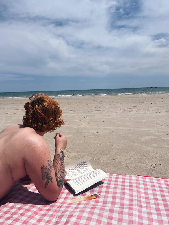 An old image of Riley, with his hair still grown out a few inches. His glasses are tilted downwards, looking at something. We see him through the gap between two bookshelves, with a dozen books in a row sat in the foreground of the image.